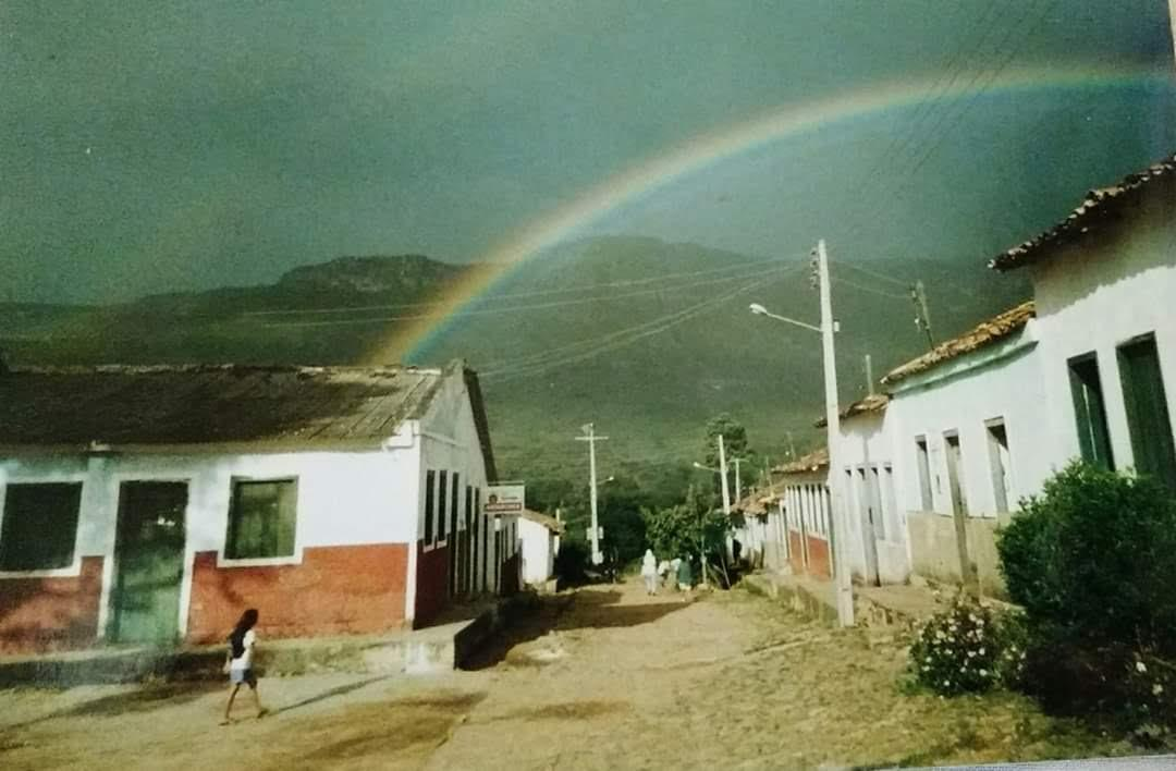Foto de época de uma rua do Vale do Capão, com um arco-íris sobre as casas e a serra ao fundo